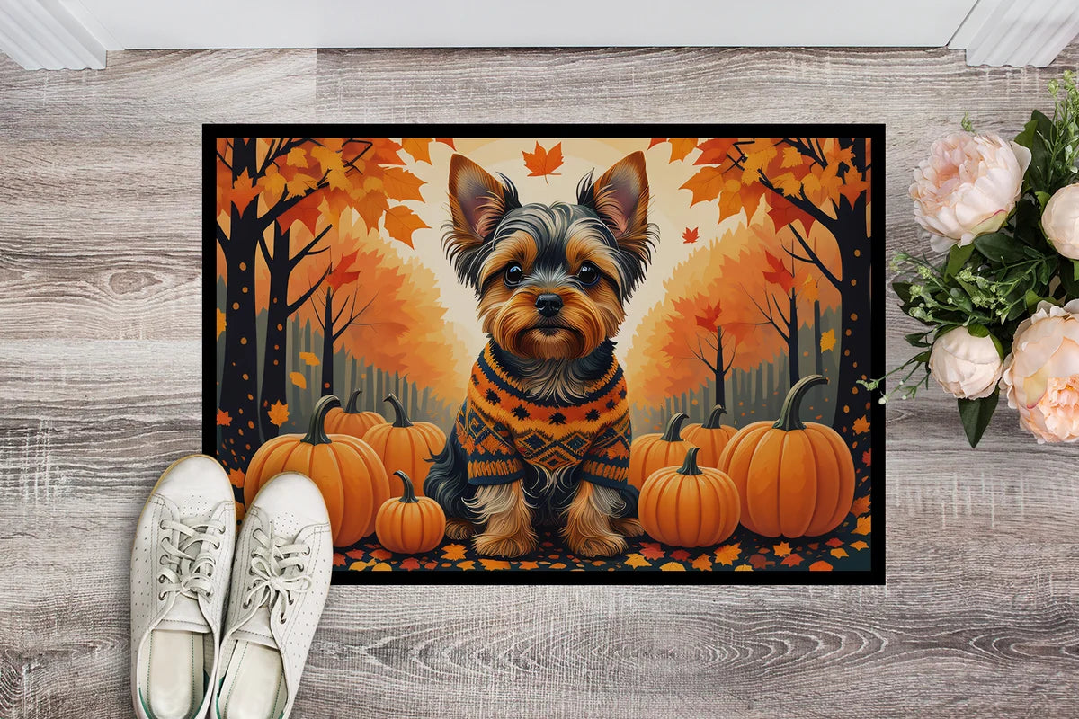 A rectangular doormat featuring a small dog in a sweater surrounded by pumpkins and autumn trees, placed on a gray wood floor with white shoes and pink flowers nearby.