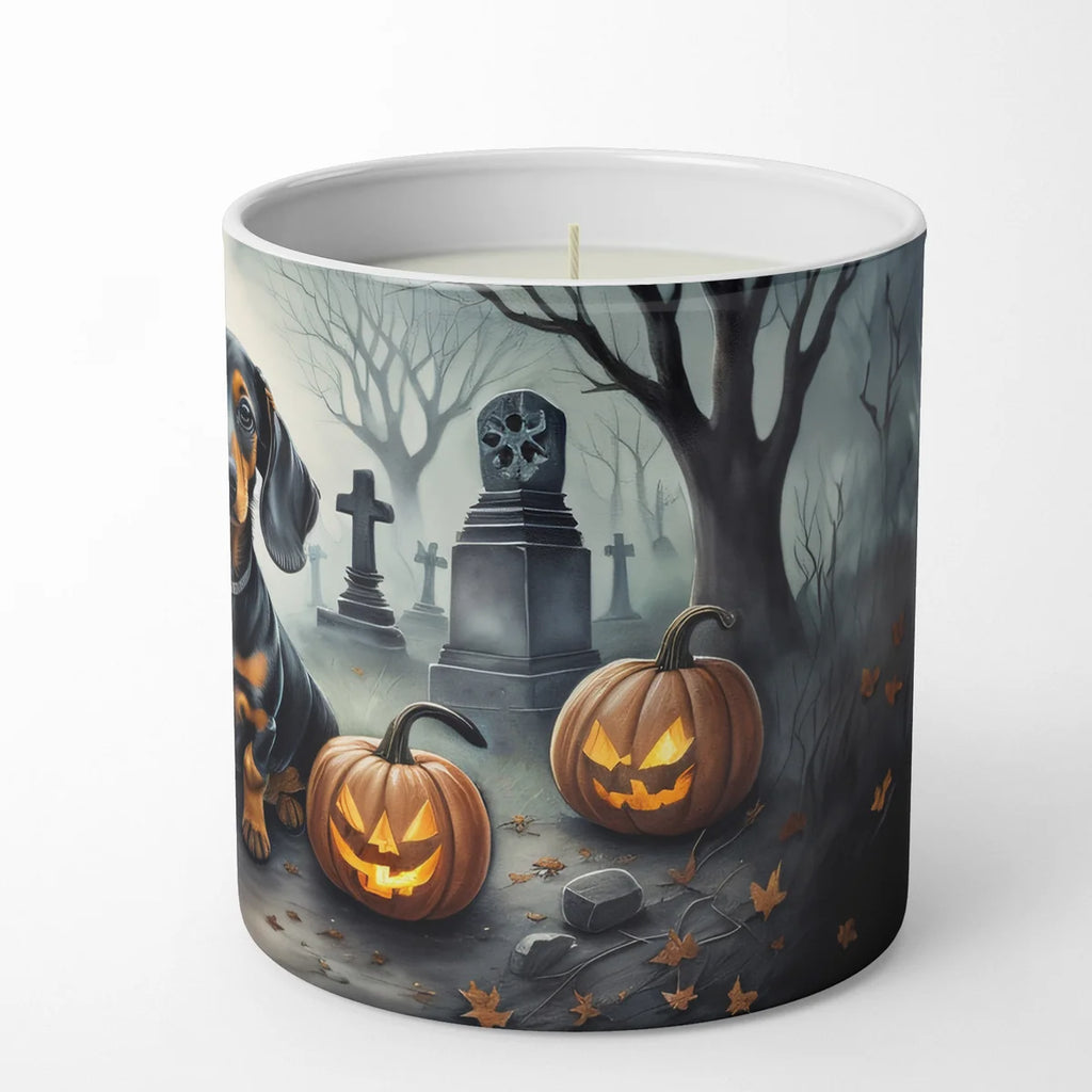 A white wax candle in a cylindrical ceramic container featuring a Halloween-themed illustration of a dachshund, tombstones, bare trees, and glowing jack-o'-lantern pumpkins on a light gray background.