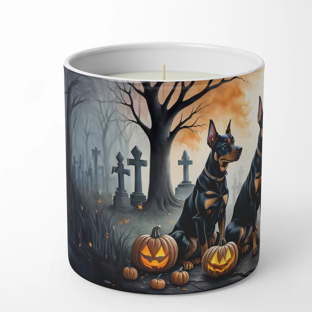 A white ceramic candle jar featuring a detailed Halloween illustration of two black and tan Doberman dogs sitting in a spooky graveyard with jack-o'-lanterns and bare trees.