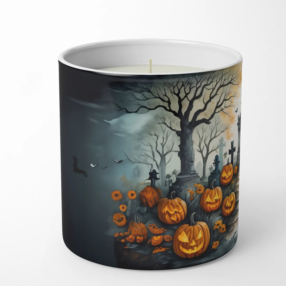 A white wax candle in a cylindrical ceramic mug with a printed Halloween scene on the outside. The artwork shows a dark graveyard with bare trees, tombstones, flying bats, and glowing jack-o'-lantern pumpkins under a cloudy night sky.