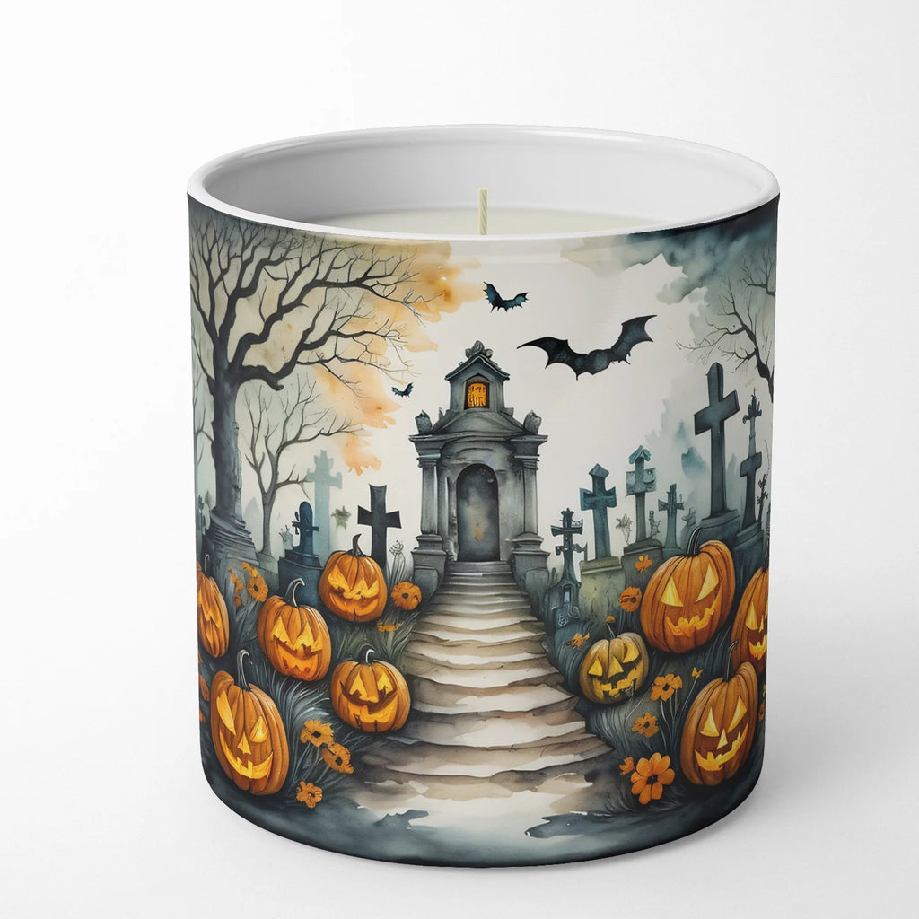 A white wax candle in a cylindrical container with a printed Halloween-themed illustration featuring a stone tombstone pathway, jack-o'-lanterns, bare trees, bats, and crosses under a cloudy sky.