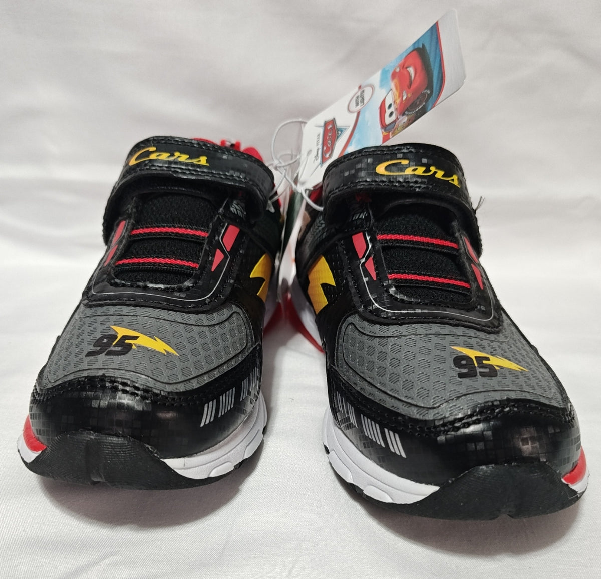 A pair of children's black and gray running shoes with yellow and red accents, featuring a hook-and-loop strap closure and a hangtag, photographed on a white fabric background.