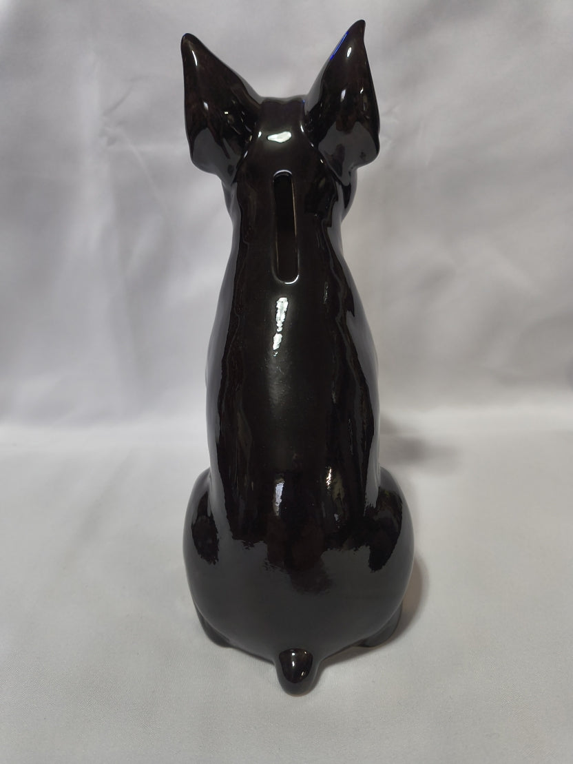 A glossy black ceramic cat bank with a coin slot cutout on its back, displayed on a wrinkled white fabric background.