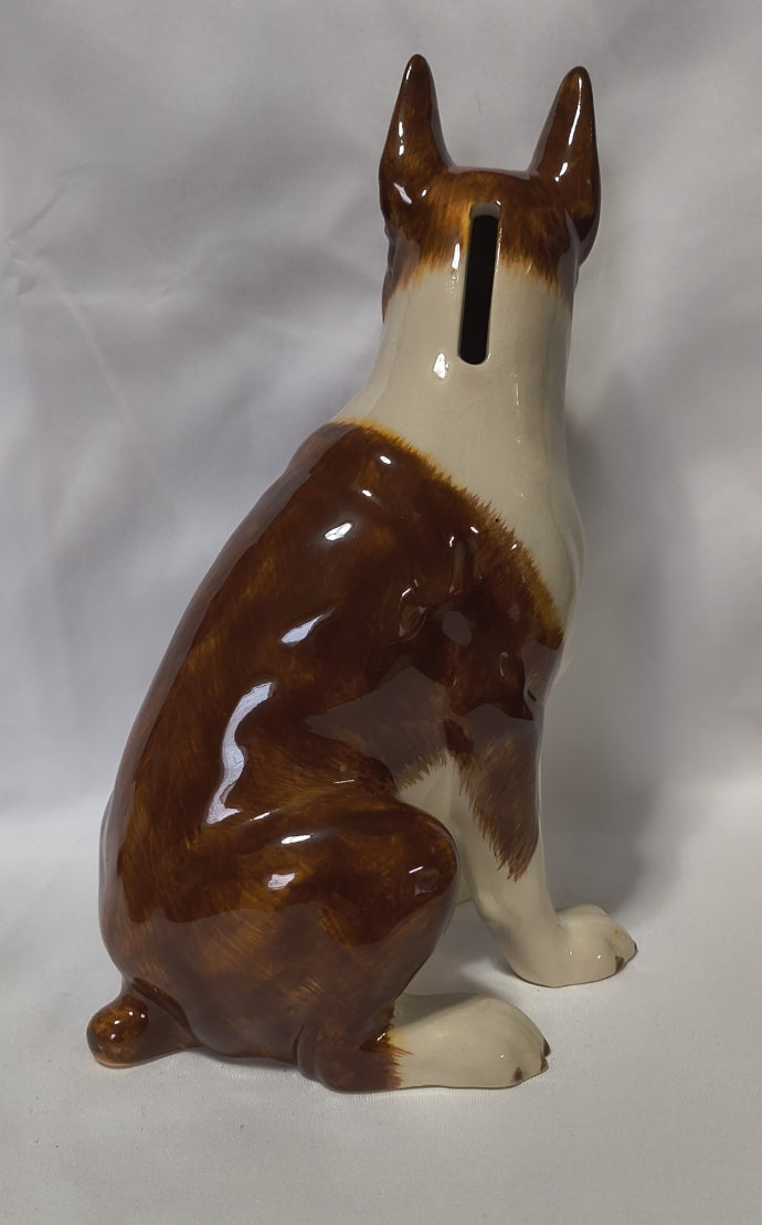 A glossy ceramic dog bank shaped like a brown and cream boxer, viewed from the back to show the vertical slot opening, set against a plain light background.