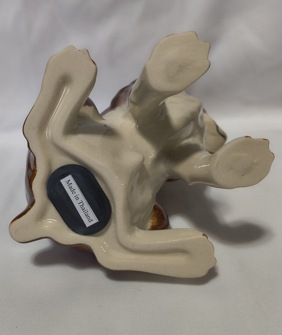 A ceramic bank of a stylized animal, a boxer dog, shown from underneath on a white background. The bank has a glossy cream-colored glazed body with brown painted details on the paws and legs, and a small black oval stopper that reads 'Made in Thailand'.