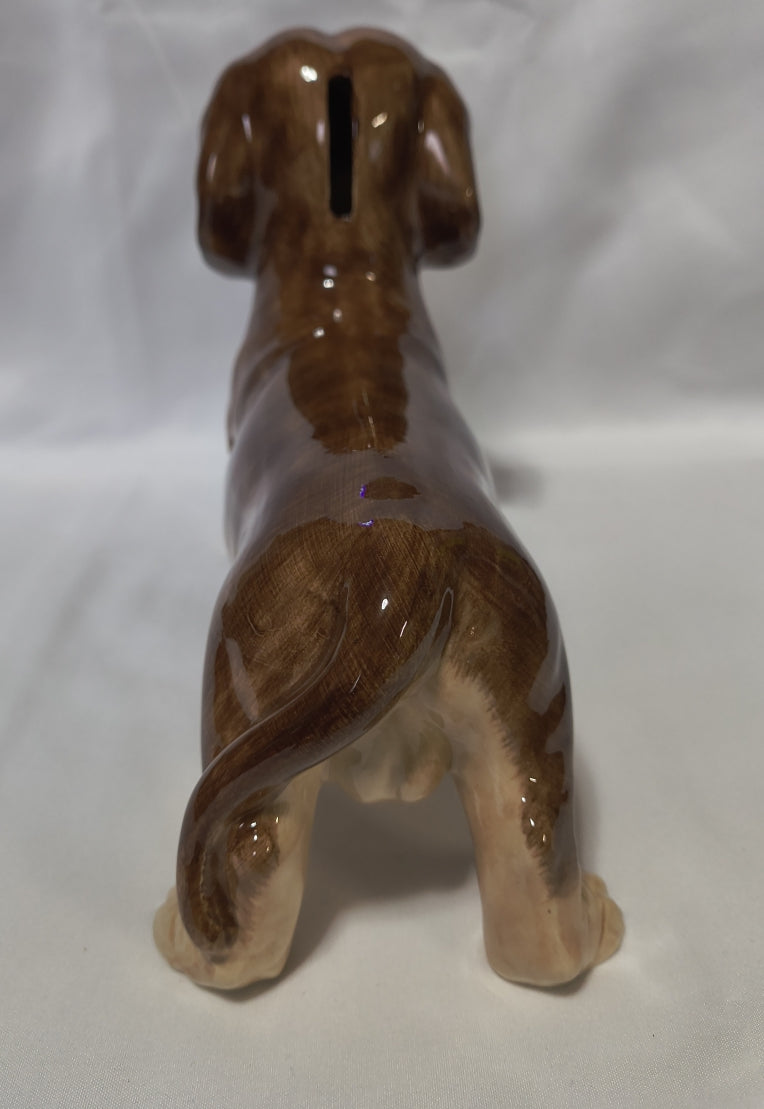 A glossy ceramic dachshund bank shown from the back, featuring a vertical slot for inserting coins, on a white fabric background.