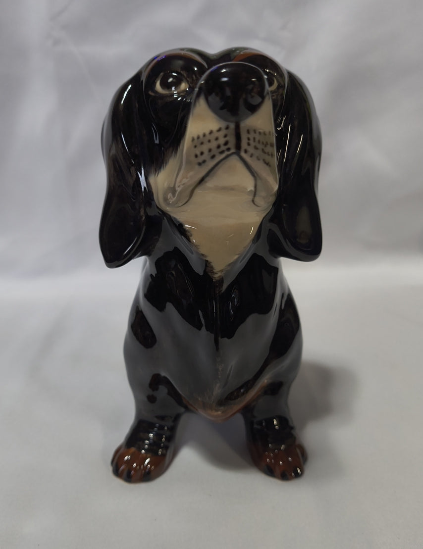 A glossy black ceramic bank of a dachshund dog, featuring a cream-colored snout and muzzle with brown paw pads and toes, set against a wrinkled white fabric background.