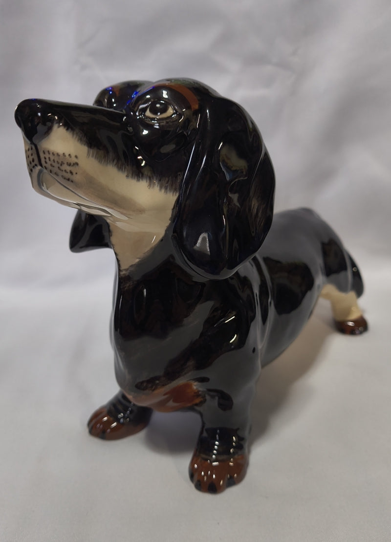 A glossy ceramic dachshund bank in black, cream, and brown, shown on a white fabric background.