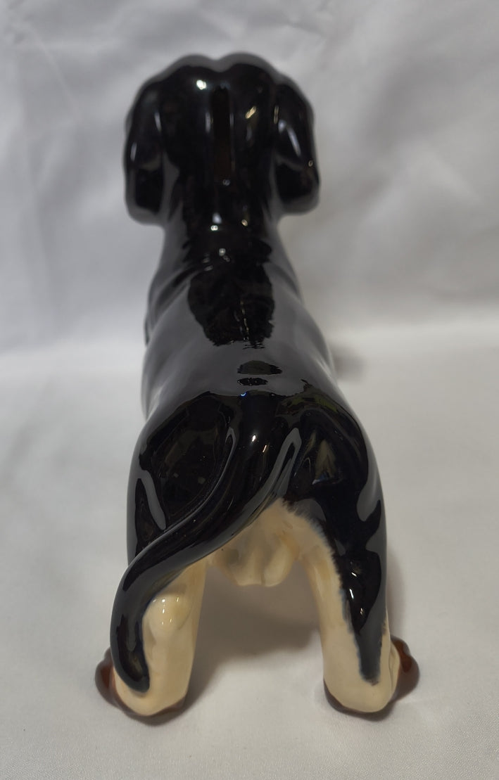 A glossy black ceramic dog bank with cream-colored paws and tail, shown from a top-down angle on a white fabric background.