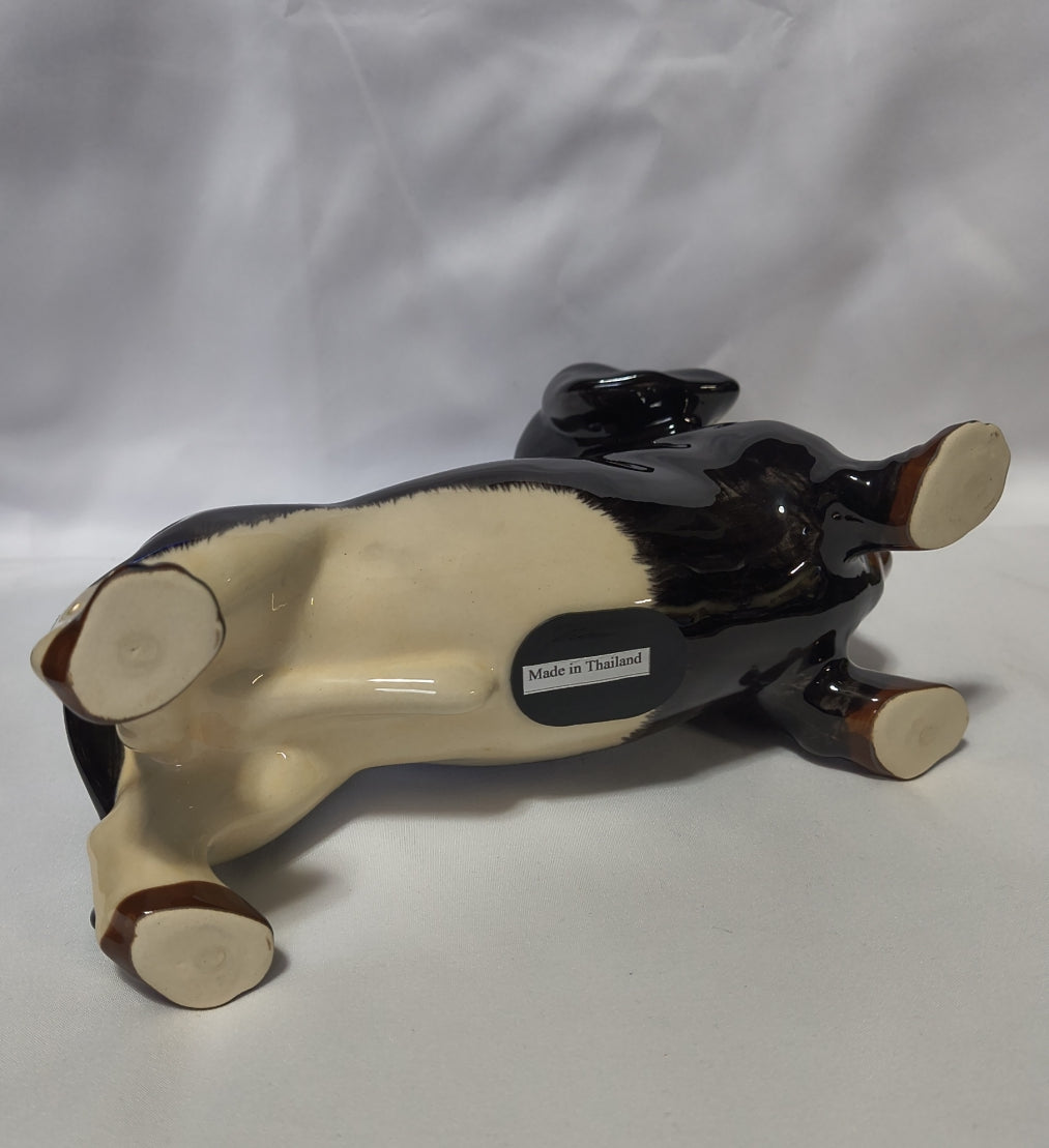 A glossy ceramic dog bank shown upside down on a white fabric background. The figurine has a black body with a cream-colored underbelly and paws.
