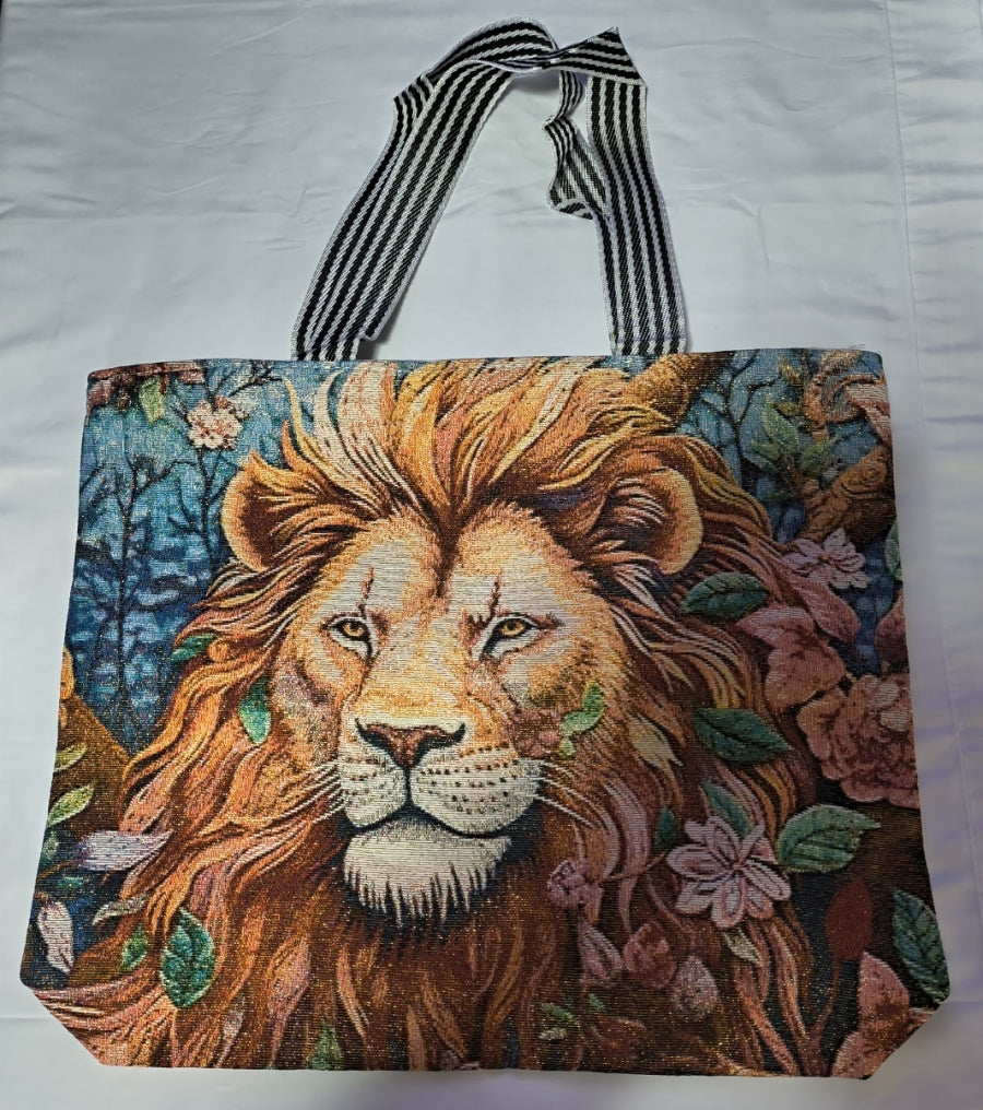 A woven tote bag featuring a detailed tapestry-style lion face surrounded by flowers and leaves, with black and white striped handles, laid flat on a white fabric background.