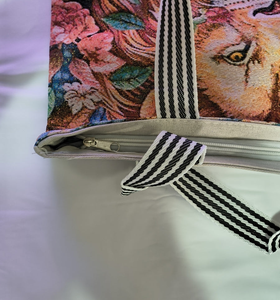 Close-up of a zippered fabric tote or document bag with a colorful floral and animal print, featuring black-and-white striped webbing handles and a matching tie detail, laid on a white surface.