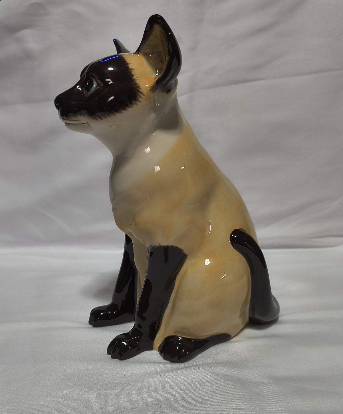 A glossy ceramic figurine of a sitting cat with a cream and black coloration, resembling a Siamese cat, set against a white fabric background.