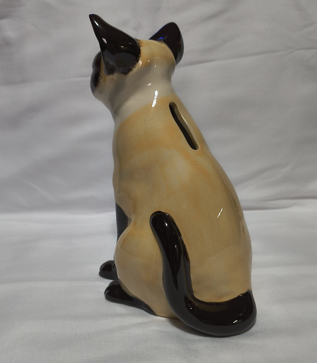 A ceramic cat-shaped pig bank shown from the back, featuring a cream-colored body with dark brown or black points, photographed against a white fabric background.