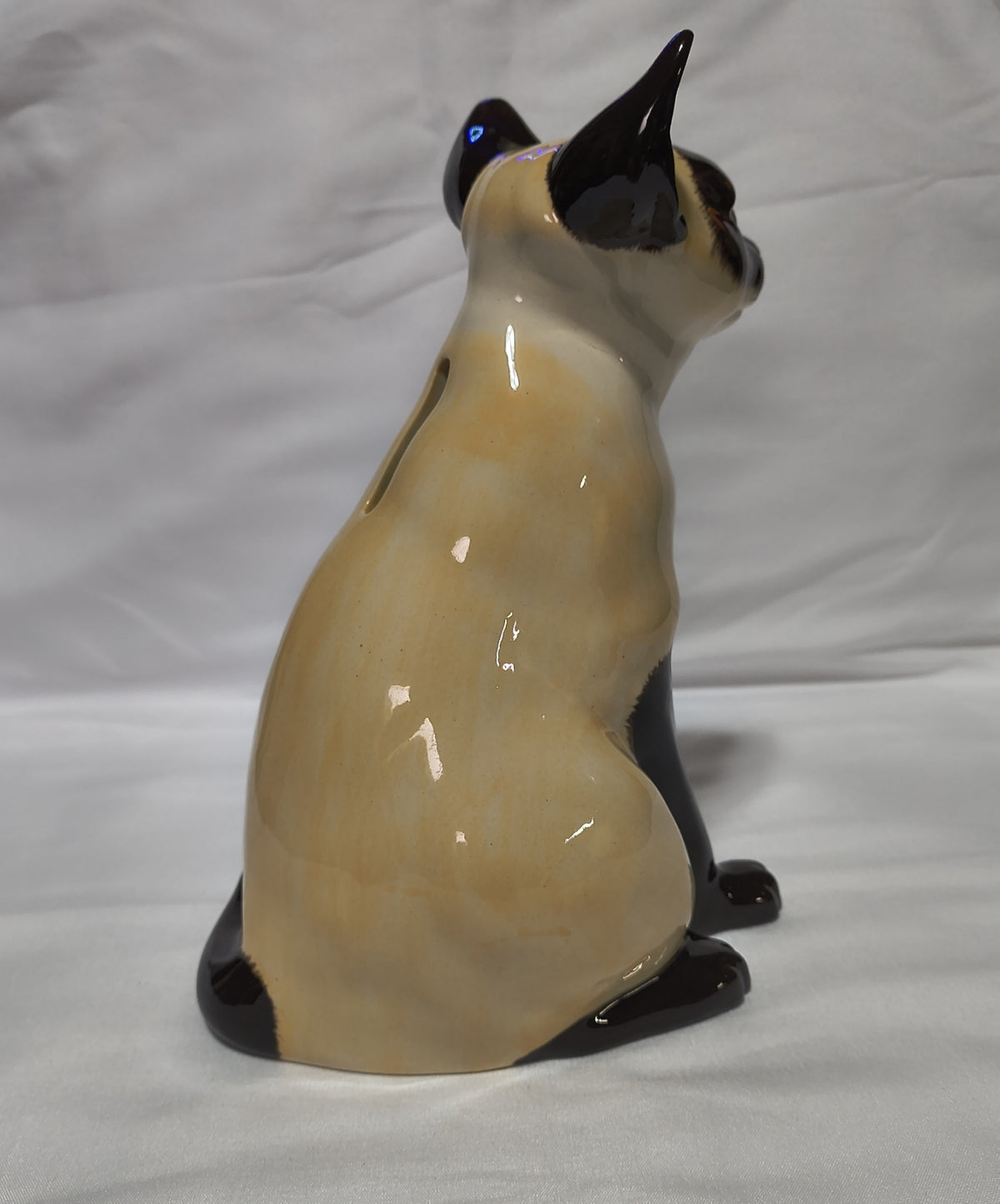 A glossy ceramic cat figurine shown from the back, featuring a cream-colored body with black paws, ears, and face, set against a wrinkled white fabric background.