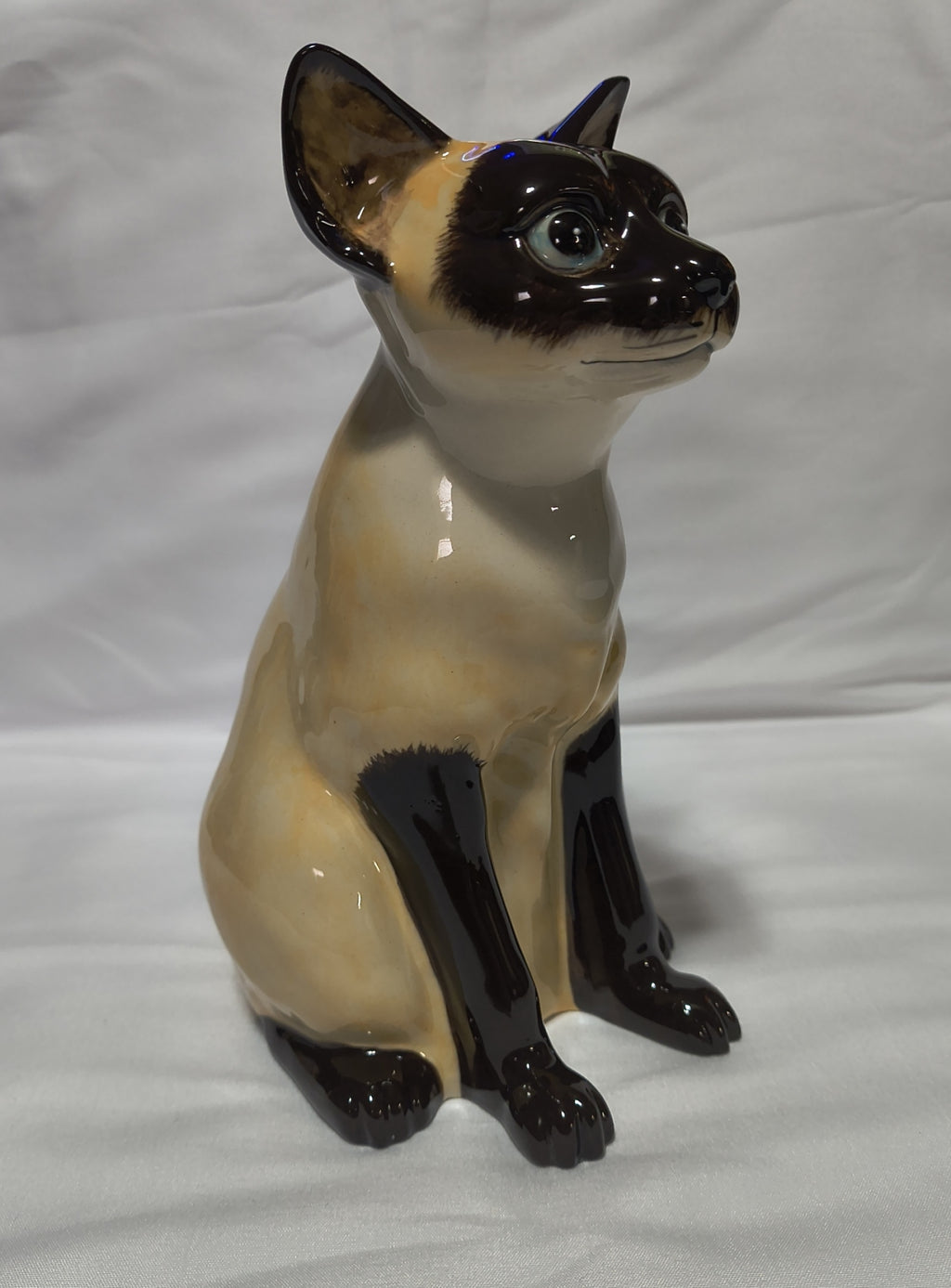A glossy ceramic figurine of a Siamese cat in a sitting position, featuring a cream-colored body with dark brown to black facial, paws, and legs, set against a white fabric background.