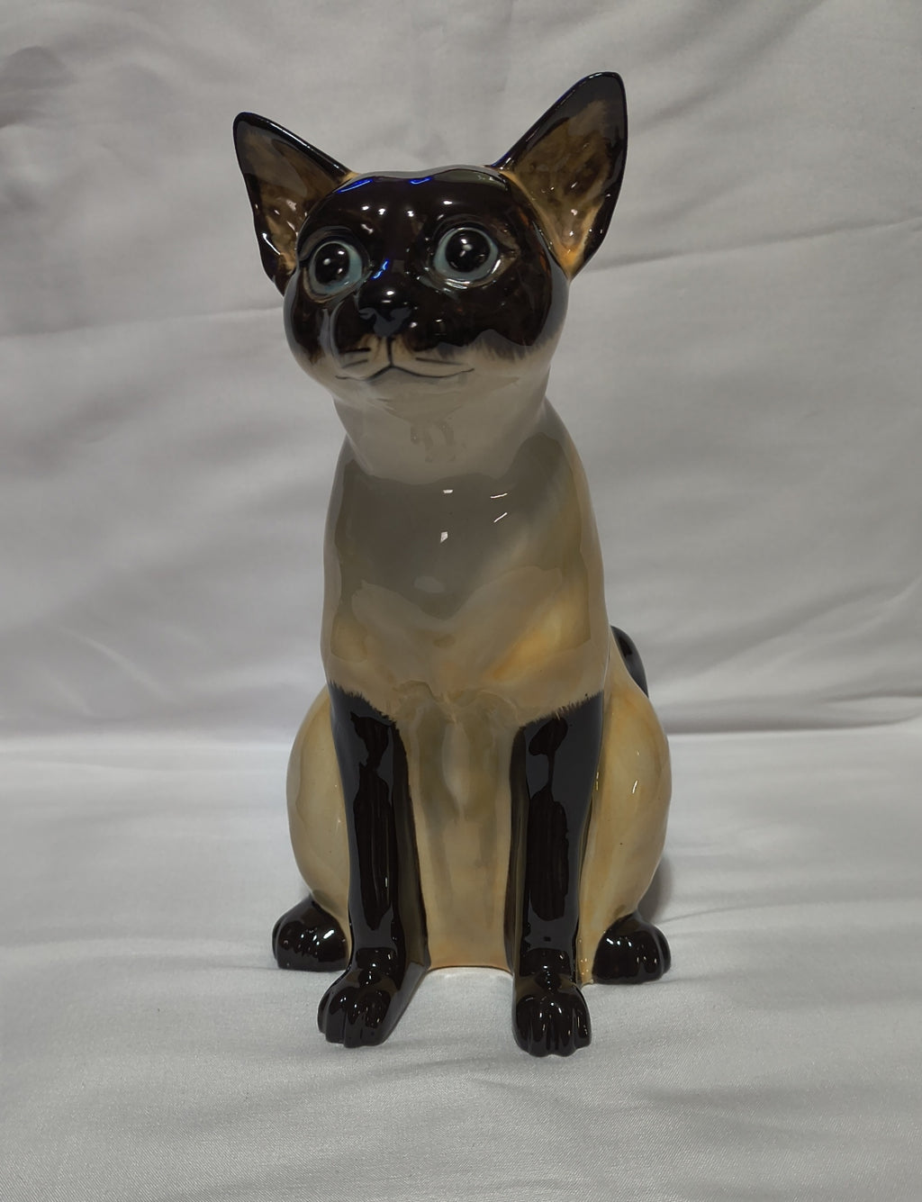 A glossy ceramic figurine of a Siamese cat sitting upright, featuring a cream-colored body with dark brown to black facial, paws, and legs, set against a plain white fabric background.