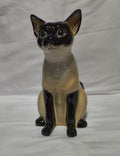 A glossy ceramic figurine of a Siamese cat sitting upright, featuring a cream-colored body with dark brown to black facial, paws, and legs, set against a plain white fabric background.
