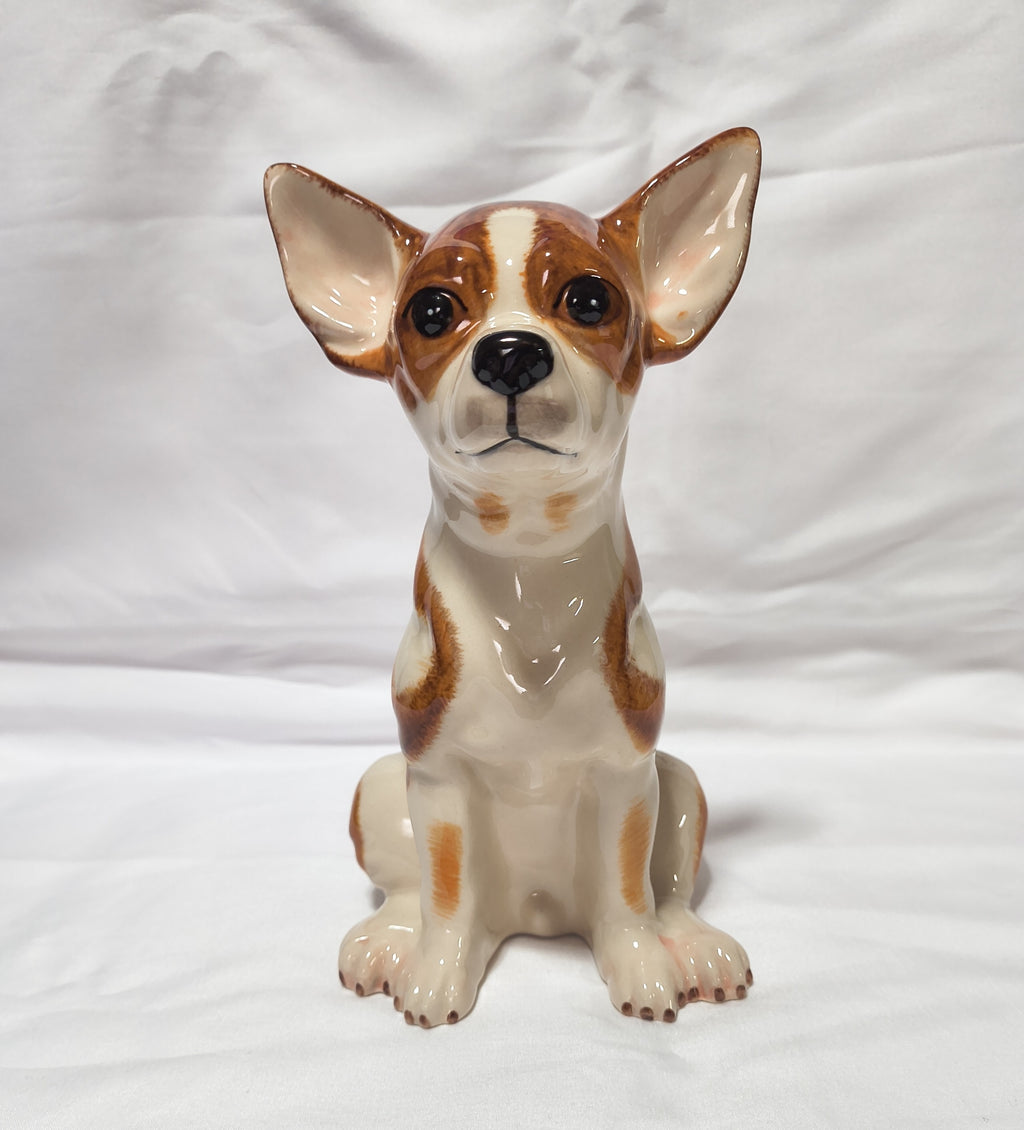 A ceramic figurine of a sitting Chihuahua dog with brown and cream coloring, photographed against a white fabric backdrop.