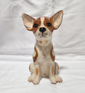 A ceramic figurine of a sitting Chihuahua dog with brown and cream coloring, photographed against a white fabric backdrop.