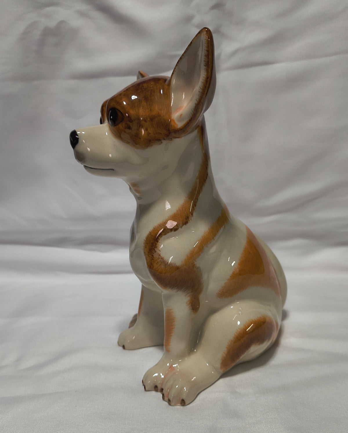 A glossy ceramic figurine of a sitting Chihuahua dog in cream and brown glaze, photographed against a wrinkled white fabric background.