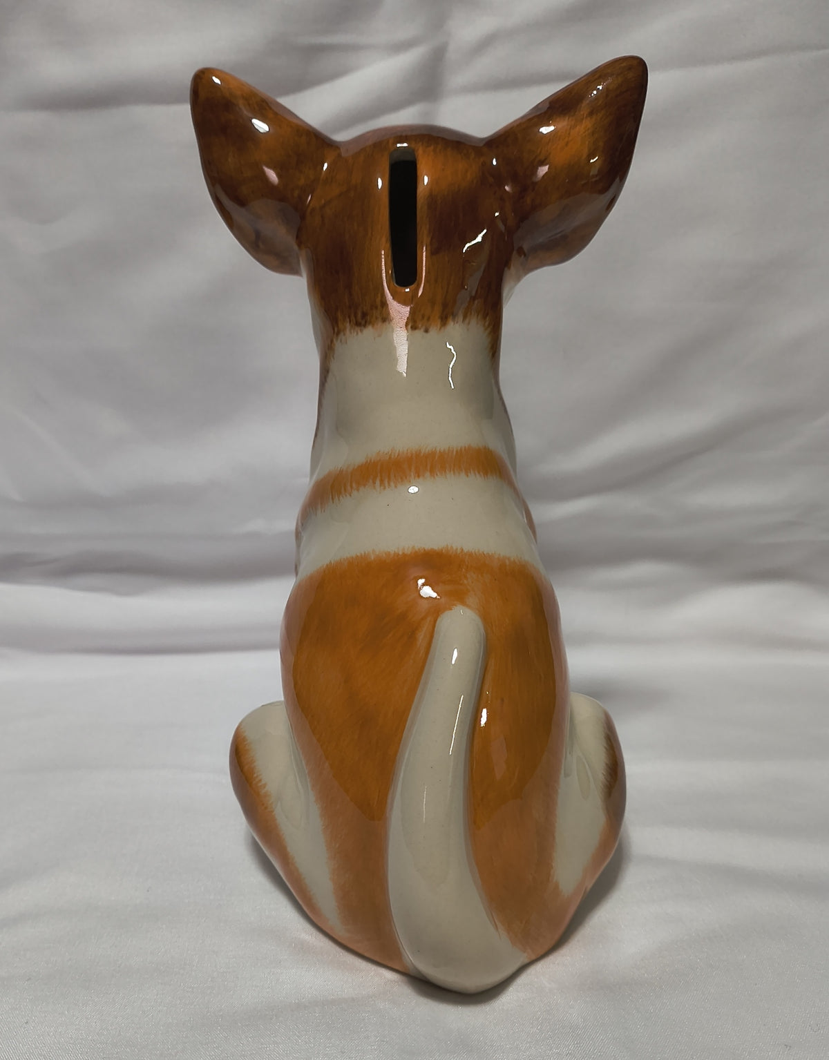 A glossy ceramic dog pig bank shaped like a sitting dog, viewed from the back. It features a brown and cream color scheme, with a vertical slot for inserting coins at the top of the ears, set against a white fabric background.