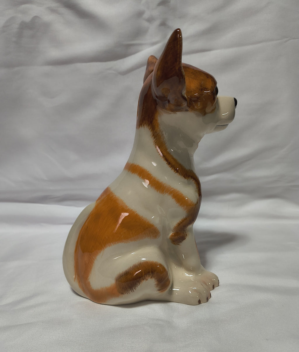 A glossy ceramic dog figurine, shaped like a sitting Chihuahua, in white and brown glaze, set against a white fabric backdrop.