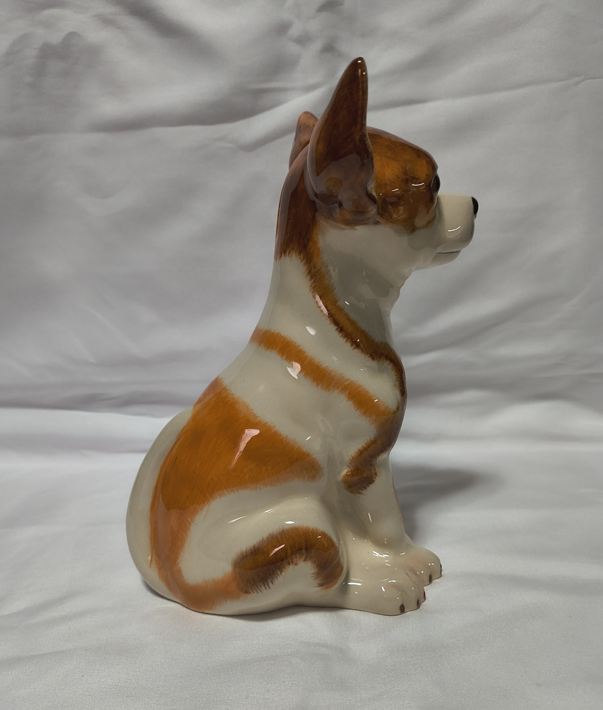 A glossy ceramic dog figurine, shaped like a sitting Chihuahua, in white and brown glaze, set against a white fabric backdrop.