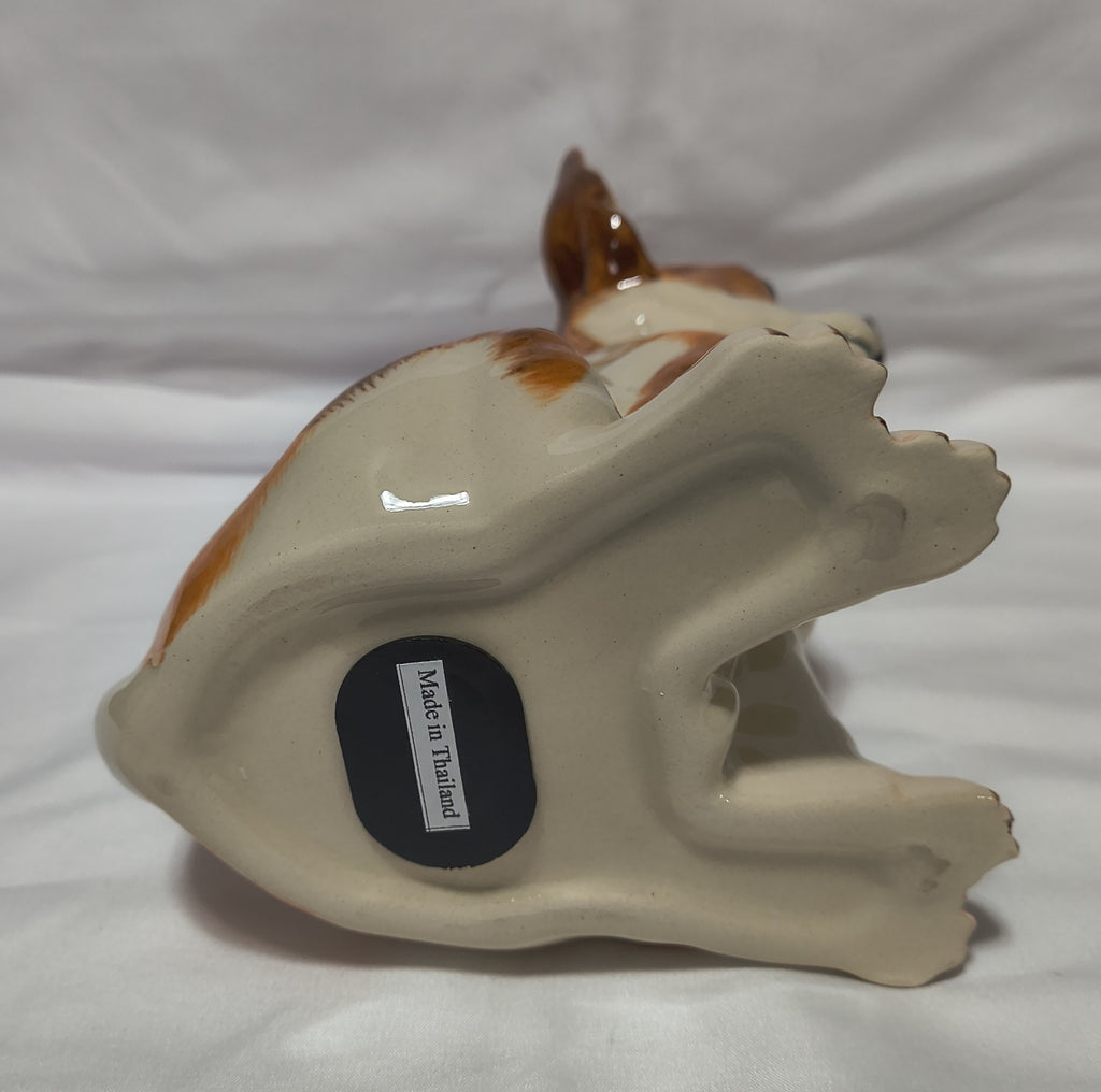A ceramic dog figurine shown from the bottom, featuring a glossy cream-colored body with brown and white patches, resting on a white fabric background. A black circular stopper on the bottom reads 'MADE IN Thailand'.