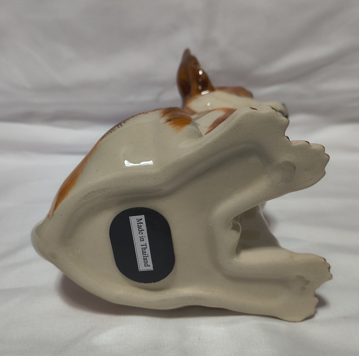 A ceramic dog figurine shown from the bottom, featuring a glossy cream-colored body with brown and white patches, resting on a white fabric background. A black circular stopper on the bottom reads 'MADE IN Thailand'.