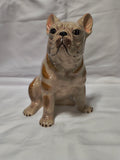 A ceramic French Bulldog figurine in a sitting position, featuring a glossy cream and tan glaze with brown markings, set against a white fabric backdrop.