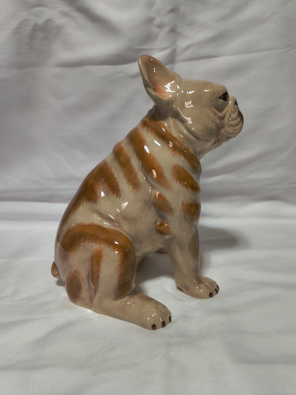 A glossy ceramic figurine of a French Bulldog in a sitting position, featuring orange and cream-colored stripes, set against a white fabric backdrop.