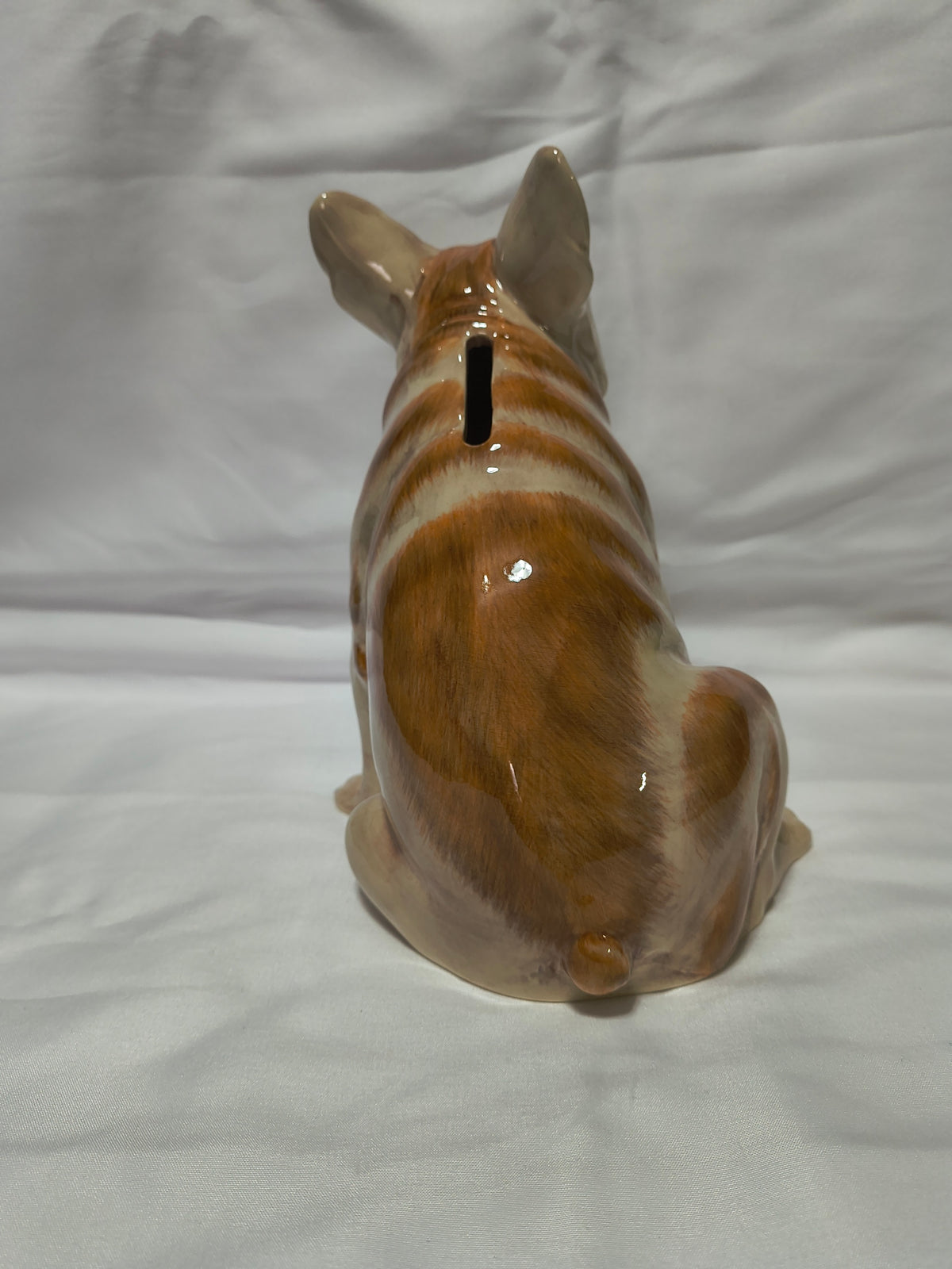 A glossy ceramic dog-shaped pig bank shown from the back, featuring brown and cream stripes with a black slot for coins, set against a wrinkled white fabric background.