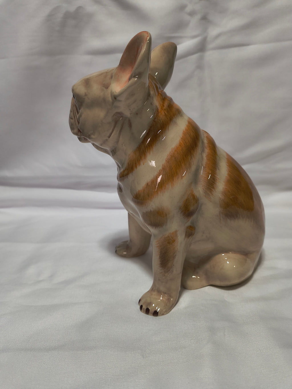 A glossy ceramic figurine of a French Bulldog in a sitting position, featuring a cream-colored glaze with orange-brown stripes, set against a white fabric backdrop.