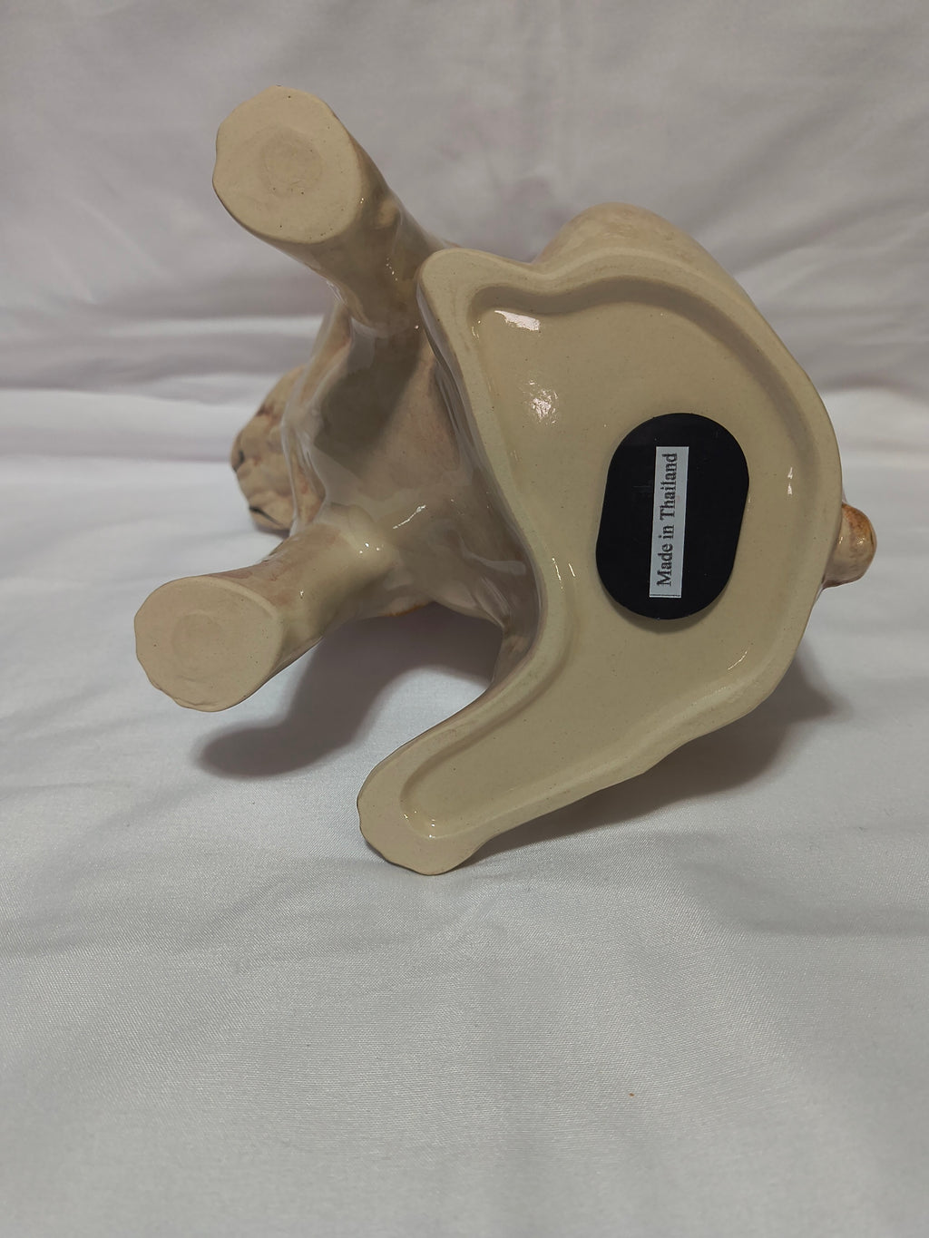 A beige ceramic figurine shown upside down on a white fabric background. A small black sticker on the bottom reads 'Made in Thailand'.