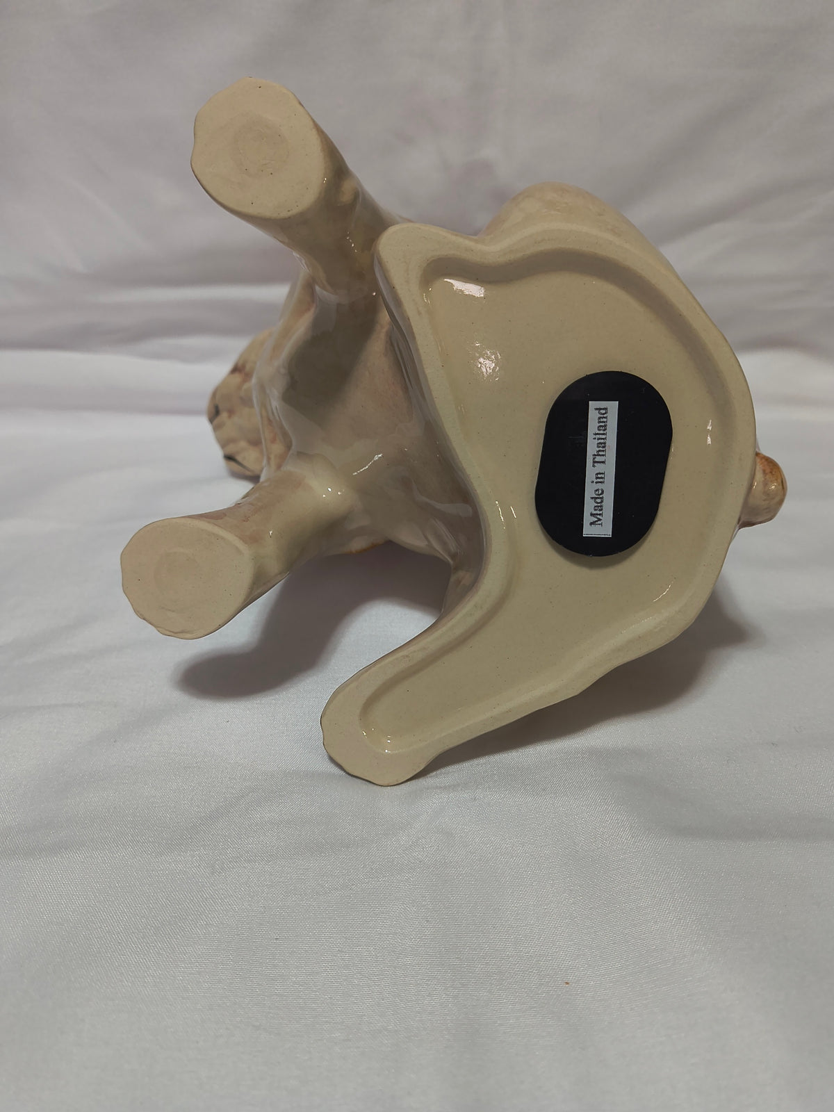 A beige ceramic figurine shown upside down on a white fabric background. A small black sticker on the bottom reads 'Made in Thailand'.