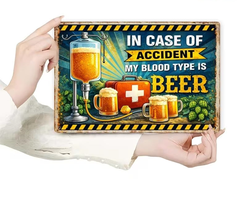 A person holds a rectangular decorative tin sign with a humorous retro-style beer sign that reads 'IN CASE OF ACCIDENT MY BLOOD TYPE IS BEER'. The sign features illustrations of an IV bag filled with amber beer, a red first-aid kit, mugs of beer, and hops on a dark teal background with yellow and black hazard stripes.