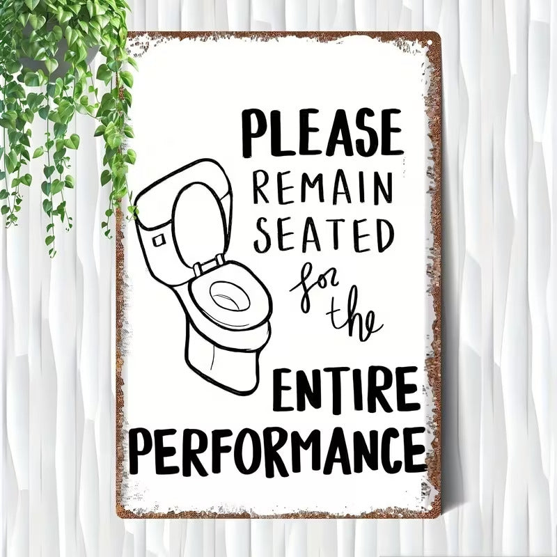 A rectangular metal sign with a distressed rusted border, featuring a black line drawing of an open toilet and the text 'PLEASE REMAIN SEATED for the ENTIRE PERFORMANCE' on a white background. The sign is displayed against a white textured wall with a green hanging plant visible in the top-left corner.